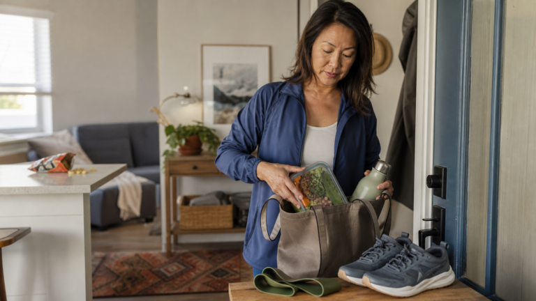 A midlife woman packs a balanced lunch and water into a work tote beside walking shoes and a resistance band before leaving home.