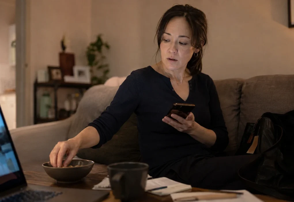 A professional woman looks at her phone while reaching automatically into a nearly empty snack bowl beside a laptop at home.