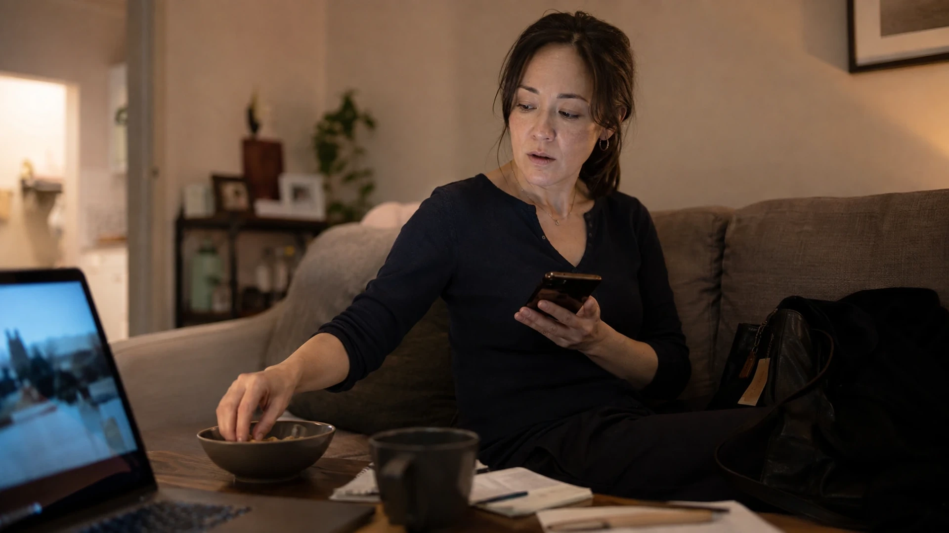 A professional woman looks at her phone while reaching automatically into a nearly empty snack bowl beside a laptop at home.